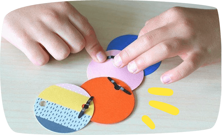 A child's hands assembling colorful paper circles together using metal brads on a light wooden surface.