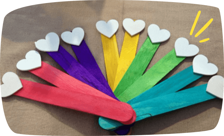 A fan of colorful painted craft sticks arranged in a rainbow pattern, each topped with a small white wooden heart.